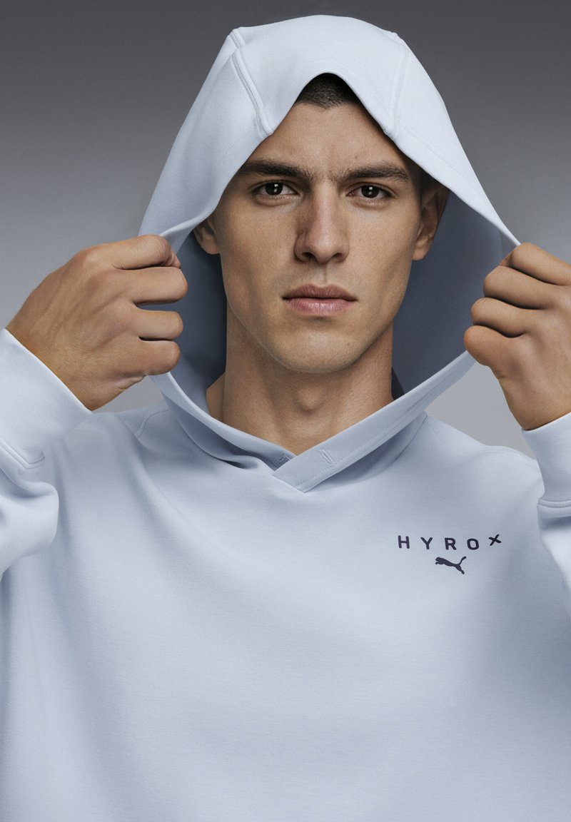 Νεαρός άνδρας που ρυθμίζει τη κουκούλα ενός ανοιχτό μπλε φούτερ Hyrox Puma, κοιτάζοντας ευθεία μπροστά με ουδέτερη έκφραση.