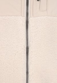 Chaqueta de forro polar con cierre de cremallera. La parte superior es lisa, de color beige claro, mientras que la parte inferior presenta una textura esponjosa de color blanco roto.