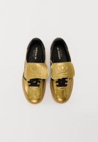 Zlaté metalické tenisky s černým jazykem a tkaničkami, vybavené texturovaným suchým zipem a hnědou gumovou podrážkou. Viditelné logo Adidas.