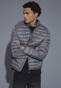 Noor mees lokkis juustega, kes kannab halli tikandiga jopet, käed ristatud, vaatab vasakule ühevärvilise halli tausta ees.