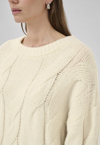 Cremfarbener Strickpullover mit strukturiertem Muster, der über einen weiten Rippkragen und offene Strickdetails an den Ärmeln verfügt.