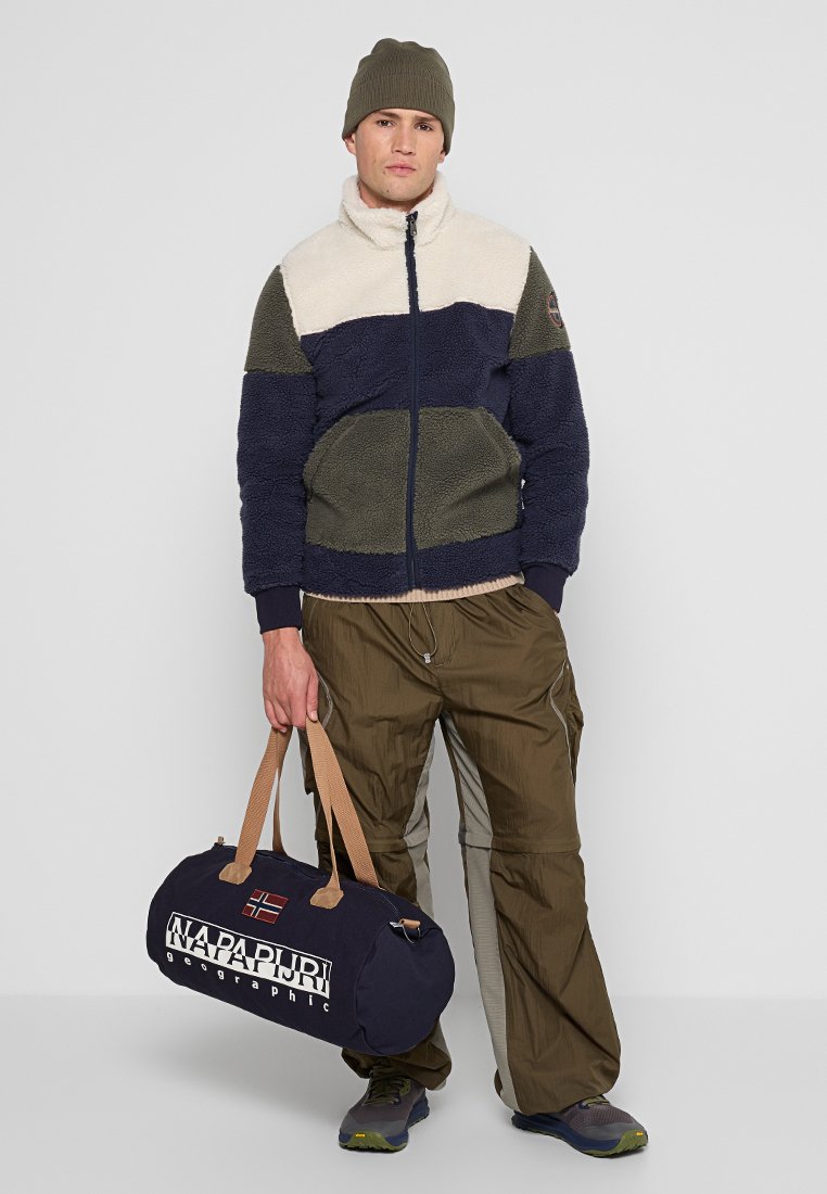 Mężczyzna w kolorowej polarowej kurtce, oliwkowych spodniach, czapce beanie i sneakersach, trzymający granatową torbę sportową z marką „Napapijri”. 