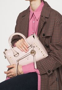 Borsa in pelle rosa chiaro con borchie dorate, manici superiori e dettagli con cerniera. Presenta un charm con logo circolare e branding testuale sulla parte anteriore.