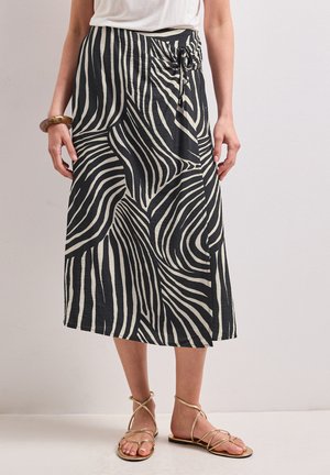 Kvinde iført en sort-hvid zebra-stribet midi-nederdel med en sløjfe i siden, kombineret med gyldne flade sandaler med stropper og en hvid top.