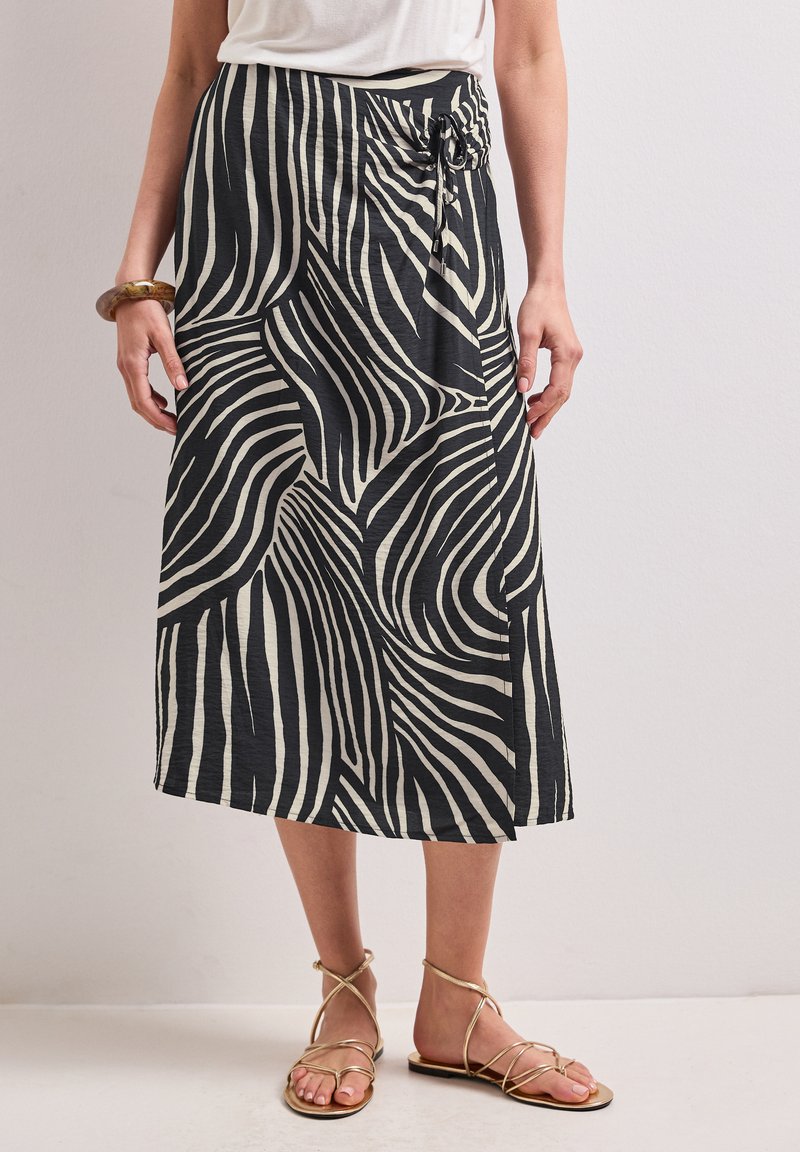 Femme portant une jupe midi à rayures zèbre noir et blanc avec un lien sur le côté, associée à des sandales plates dorées à brides et un top blanc.