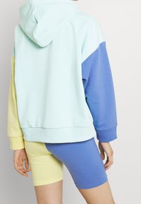 Persona vestida con una sudadera de bloques de color con mangas en azul pálido, amarillo y azul, combinada con pantalones cortos de ciclismo a rayas en amarillo y azul.