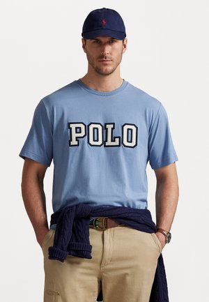 Tricou din bumbac de culoare albastru deschis, cu inscripție îndrăzneață "POLO", purtat cu pantaloni khaki și un pulover închiriat de culoare bleumarin în jurul taliei. Șapcă bleumarin expusă.