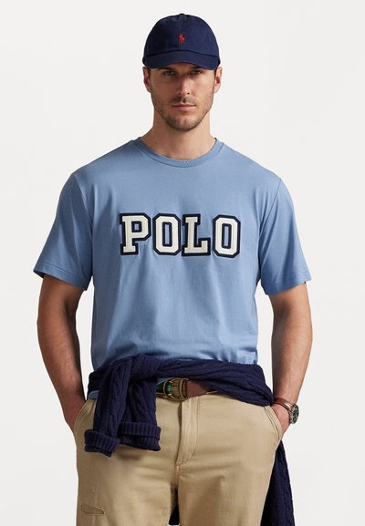 T-shirt en coton bleu clair avec l'inscription "POLO" en lettres bold, porté avec un pantalon kaki et un pull navy foncé drapé autour de la taille. Casquette navy présentée.