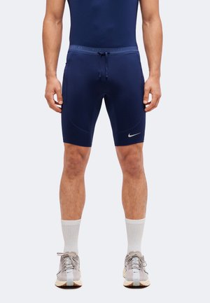 Homme portant un short de sport bleu marine avec un cordon de serrage et un logo Nike blanc, des chaussettes blanches et des chaussures de course grises, debout droit.