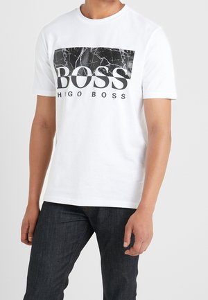 Mann trägt weißes kurzärmeliges T-Shirt mit schwarzer Marmorgrafik und "BOSS HUGO BOSS" Text auf der Vorderseite, kombiniert mit dunklen Jeans.