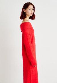 Abito lavorato a maglia rosso, off-the-shoulder, con maniche lunghe, caratterizzato da una scollatura a costine testurizzata e una silhouette dritta fino al pavimento.