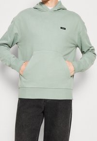 Sudadera verde menta hecha de una tela suave, con un bolsillo frontal, puños acanalados y una pequeña etiqueta de marca en el pecho.