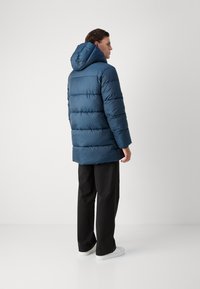 Giacca a vento blu scuro con cappuccio, caratterizzata da un design trapuntato. Abbinata a pantaloni neri e scarpe da ginnastica bianche, vista da dietro.
