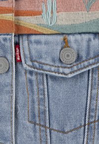 Ljusblå denimtyg med kontrasterande orange sömmar. Har en silverknapp med präglad logotyp och en röd etikett nära fickan. Detaljerad broderi ovan.