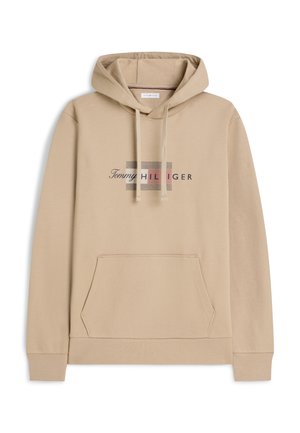 Beige hoodie with front pocket, drawstring hood, and "Tommy Hilfiger" logo printed in the center with a patterned background.