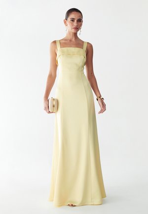 Frau in einem ärmellosen, blassgelben bodenlangen Kleid, hält eine geflochtene Clutch in der linken Hand, accessoriert mit goldenen Ohrringen und Armband.