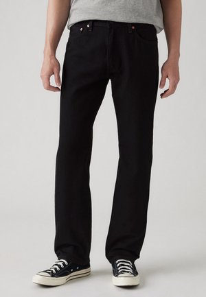 Man die zwarte jeans met rechte pijpen en zwarte lage sneakers met witte veters en zolen draagt, staand tegen een effen achtergrond.