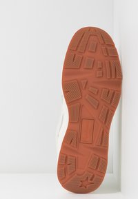 Podeszwa buta z pomarańczowej gumy o teksturowanej powierzchni, charakteryzująca się wzorem w zygzak, prostokątnymi segmentami i wytłoczonym logo. Widoczna biała górna krawędź.