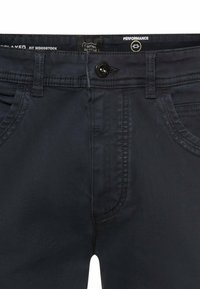 Close-up van de taille van een zwarte casual broek met knoop, riemlussen, twee voorzakken en een zichtbaar binnenlabel met merk- en pasvormdetails.