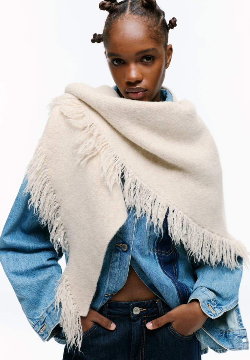 Sciarpa in lana beige con bordi frangiati appoggiata su una giacca in denim blu. Presenta una texture morbida, forma triangolare e vestibilità ampia.