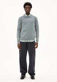 Lichtblauwe gebreide trui met ribbelboord aan de hals en manchetten, gelaagd over een lichtgroen shirt, gecombineerd met donkere wijduitlopende broeken en zwarte schoenen.