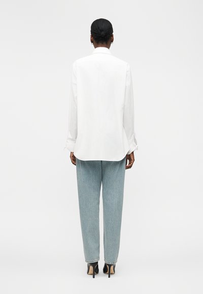 Personne aux cheveux foncés en chignon, portant une chemise blanche à manches longues, un jean bleu clair et des chaussures noires à talons hauts, debout de dos.