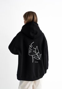Felpa nera in cotone, con un disegno lineare bianco di un volto e foglie sul retro. Include un grande cappuccio e una vestibilità ampia.