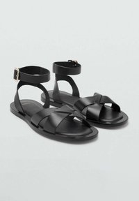 Sandali in pelle nera con più cinturini intrecciati e una fibbia alla caviglia. Suola piatta, design minimalista, texture liscia e senza motivi.