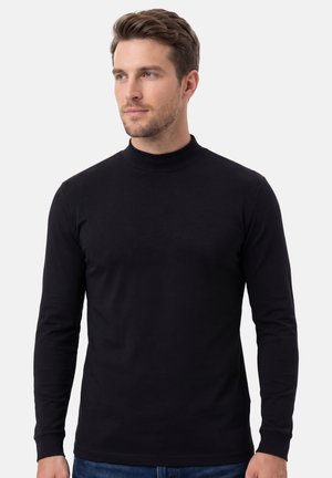 Homme aux cheveux bruns courts portant un col roulé noir ajusté à manches longues, debout devant un fond gris clair uni.