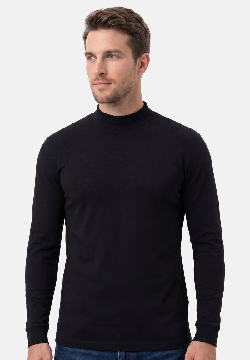 Homme aux cheveux bruns courts portant un col roulé noir ajusté à manches longues, debout devant un fond gris clair uni.