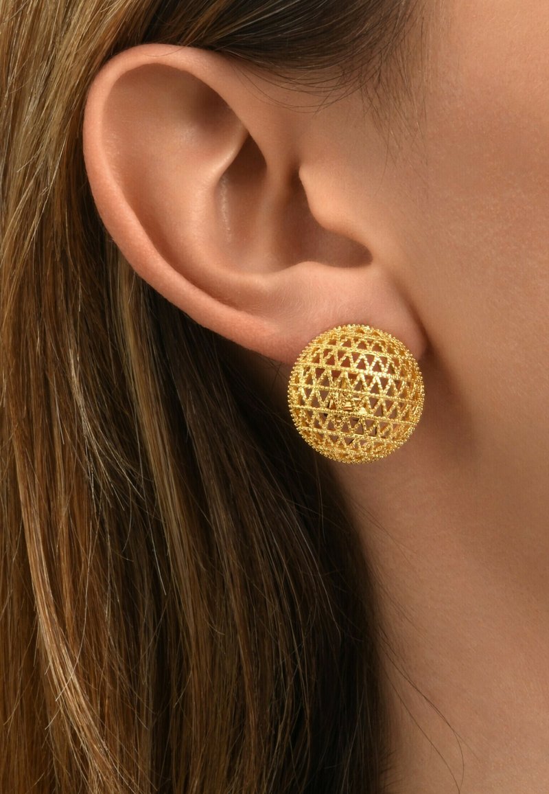 Gros plan d'une oreille portant une boucle d'oreille en acier doré à motif en treillis avec de longs cheveux bruns visibles en arrière-plan.