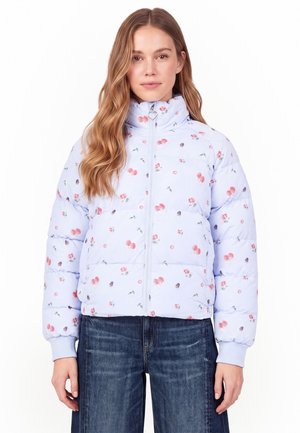 Vrouw draagt lichtblauw bloemen gewatteerd jack met rits met hartvormige trekker, gecombineerd met donkerblauwe jeans, gezicht naar voren.