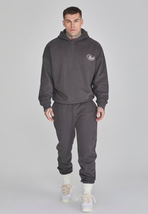 Donkergrijze, hoodie en bijpassende joggers van zachte stof. Voorzien van ribgebreide manchetten en tailleband. Witte logo op de borst. Witte sneakers.