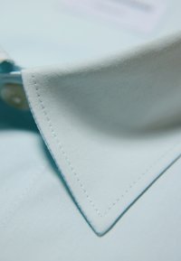 Hemelsblauwe katoenen overhemdkraag met puntige uiteinden, voorzien van fijne stiksels en een knoopdetail. Gladde textuur met een frisse uitstraling.