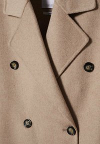 Abrigo de lana beige con diseño de doble botonadura, que presenta botones negros, solapas en forma de muesca y un acabado suave y texturizado.