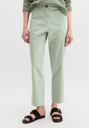 Personne portant un pantalon vert clair à jambes droites, un pull vert texturé et des sandales noires à bouts ouverts avec des boucles dorées, debout sur un fond blanc.