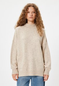 Pull beige surdimensionné avec un col montant, des manches longues et des poignets côtelés. Comprend une poche avant et un tissu doux et texturé.