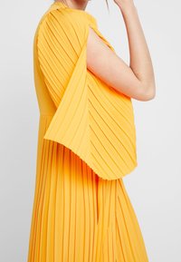 Keltainen laskostettu mekko, jossa on teksturoitu kangas, lyhyet hihat ja virtaava siluetti. Materiaali on kevyttä ja sileää.