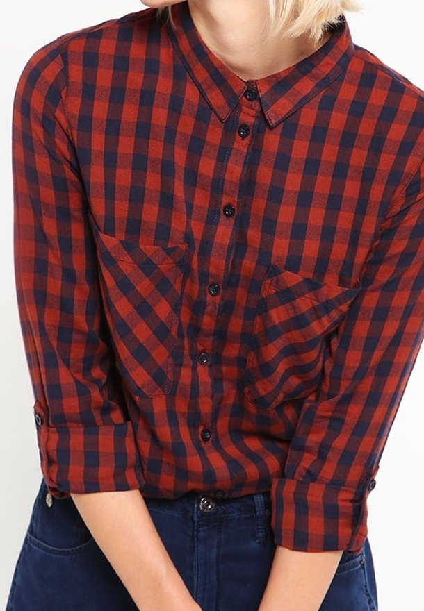 Femme portant une chemise à carreaux rouge et bleu marine à manches longues avec les manches retroussées et un pantalon bleu marine, les bras croisés à la taille.