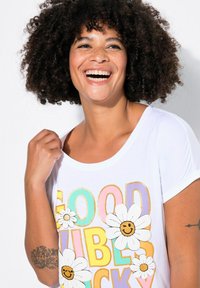 T-shirt blanc à manches courtes avec un imprimé coloré portant le texte "BONS VIBES" et des fleurs souriantes. Tissu doux avec une coupe décontractée.