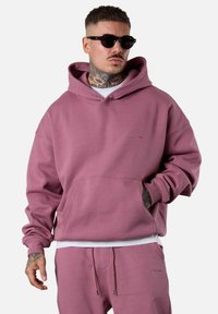 Sweatshirt à capuche rose poussiéreux en tissu doux, doté d'une poche avant et de poignets côtelés. Porté avec un jogging assorti et des lunettes de soleil.