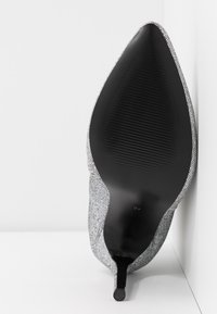 Zapato de tacón alto con parte superior de tela texturada en gris, diseño de punta afilada y suela negra acanalada. El tacón es delgado y de estilo stiletto.