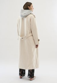 Trench coat bej cu căptușeală gri pentru glugă, talie cu curea și manșete cu nasturi. Modelul poartă pantaloni din dantelă neagră transparentă și pantofi cu vârf deschis.