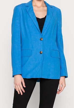 Frau trägt ein knallblaues Blazer mit zwei Knöpfen über einem schwarzen Oberteil und einer schwarzen Hose, steht vor einem schlichten Hintergrund.