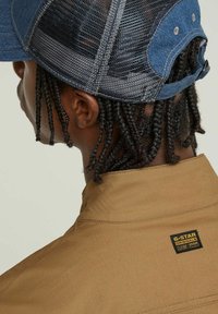 Brun jacka med hög krage och G-Star-märket, tillsammans med en blå denimkeps med nätbak och justerbar rem.