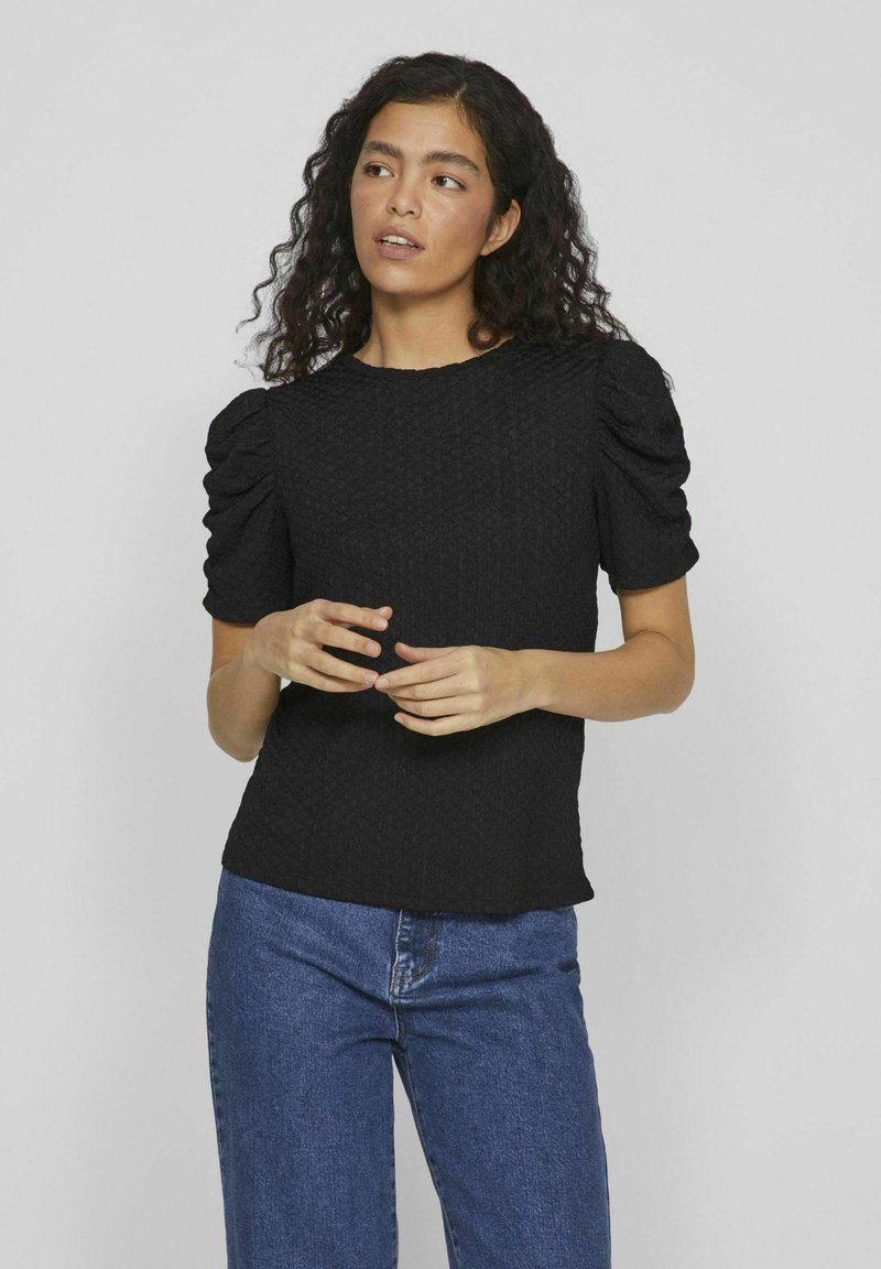 Čierna textúrovaná blúzka s nafúknutými rukávmi, okrúhlym výstrihom a slabo priliehavým strihom, skombinovaná s modrými džínami.