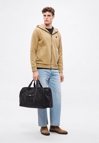 Čierna cestovná taška s dvoma hornými rúčkami, sprevádzaná opálenou mikinou na zips, svetlomodrými džínsami a hnedými členkovými topánkami.