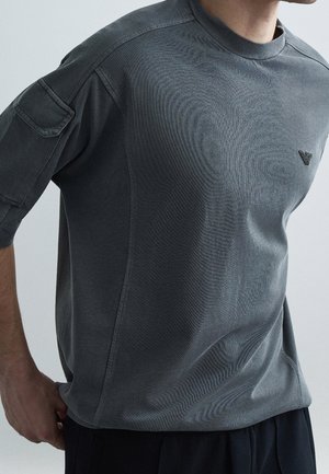 Mann trägt ein dunkelgraues kurzärmeliges Hemd mit einem Logo auf der Brust und einer Tasche am linken Ärmel vor einfarbigem Hintergrund.