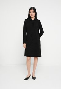 Robe noire à manches longues, col polo et cinq boutons. Présente un imprimé zèbre texturé. Portée avec des chaussures noires à talons.