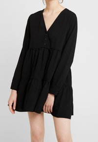Schwarzes langärmeliges Kleid mit V-Ausschnitt, geknöpftem Front und gestuftem Rüschensaum. Hergestellt aus leichtem Stoff mit einer glatten Textur.